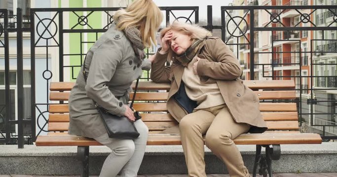Senior Caucasian woman coming to elegant retiree having headache. Ill pensioner got bad on city street. Friend worries about mature lady sitting on bench. Cinema 4k ProRes HQ.