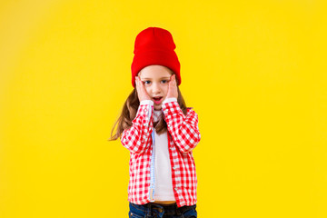 Beautiful stylish little girl dressed in pink checkered shirt, red cap and jeans on yellow orange background. Surprised shocked cute child put hand to cheek. Emotional portrait concept.