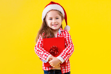 Pretty fair haired girl is holding gift box, wrapped on red paper and bow. Cute child dressed in checkered pink shirt, hat of Santa on yellow background. Emotional portrait concept. Merry christmas.