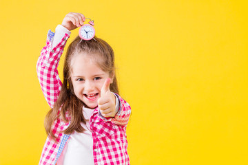 Pretty beautiful fair haired girl dressed in pink checkered shirt on yellow orange background. Cute smiling child is holding in hand little alarm clock on her head. Emotional portrait concept.