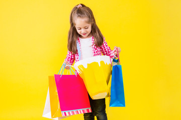 Pretty fair haired girl is holding in hands colorful bright paper bags, packages. Cute stylish child dressed in checkered pink shirt on yellow orange background. Shopping and sale concept.