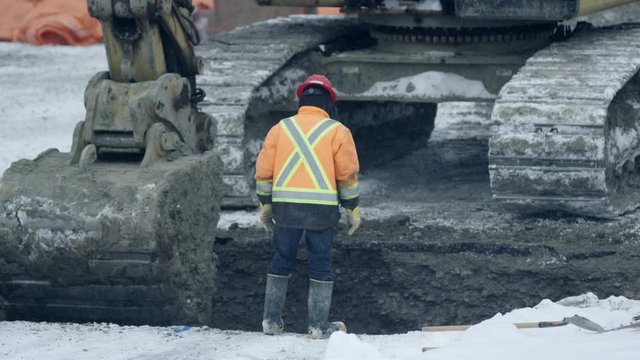 Construction workers guide excavator bucket digging hole in winter construction site on snowy day - from behind