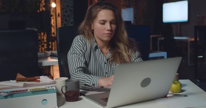 Young businesswoman spinning around at chair and started typing on her laptop. Female office worker using computer while working lon hours at empty night office. Concept of workaholism.