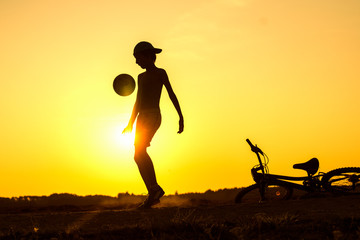 Boy playing with ball in nature, bicycle lies nearby, silhouette of playing child at sunset in countryside