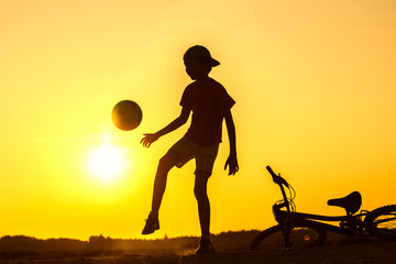 Obraz premium Boy playing with ball in nature, bicycle lies nearby, silhouette of playing child at sunset in countryside