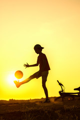 Boy playing with ball in nature, bicycle lies nearby, silhouette of playing child at sunset in countryside