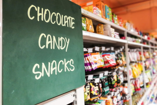 Chocolate, Candy, Snakcs Signage At The Aisle Of Supermarket With Defocused Merchandise