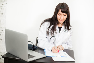 Female doctor writes on a clipboard, sitting at a desk with a laptop, white background