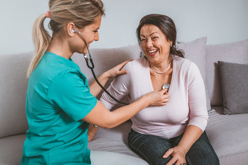 Selective focus on a serene retired woman sitting on a sofa while a female nurse using her stethoscope and listening to her lungs.