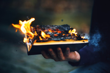 The sorcerer's hand holds a thick hardcover magic book that burns with a bright fire in the middle...
