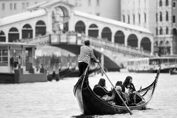 Fototapeta na wymiar Iconic black and white view of a gondolier rowing on the Grand Canal, with the Rialto bridge and a water-bus in the distance