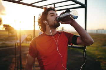Close-up portrait of a caucasian young man with earphones in his ears drinking water after exercising in the outdoor gym park - Powered by Adobe