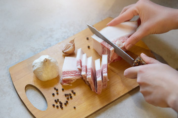 Faceless portrait of woman cutting lard with layers of meat on wooden cutting board among some garlic, peppercorns on white table, females's hands cuts fat, lady preparing snack for her husband.