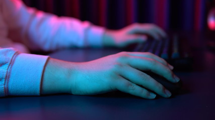 A young man types on a computer keyboard and works with a mouse. Hands close up. Blue and red light falls on the hands.