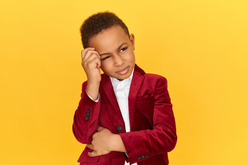 Pensive adorable little boy of African appearance looking upward keeping hand on his forehead...