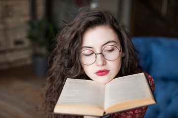 Close up portrait of attractive student girl with curly hair, in round glasses. Young woman sitting and reading book in hands near face, place her chin on it. Education and knowledge concept.