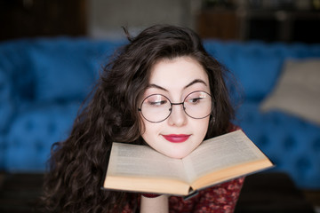 Close up portrait of attractive student girl with curly hair in round glasses. Young woman reading book in hands near face, place her chin on it. Her eyebrows and lips express skepticism and disbelief