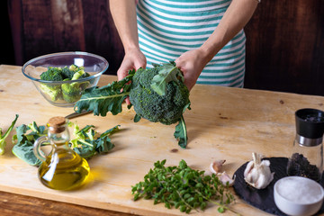 Moments of everyday life in Mediterranean cuisine: young female cook cleans broccoli on a wooden chopping board with extra virgin olive oil, garlic, pepper and knife on top