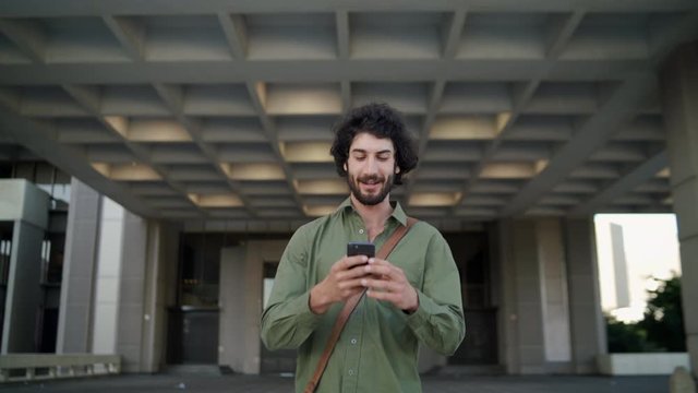 Smiling Professional Young Executive Man Come Out Of The Corporate Building Using His Smart Phone Walking Toward Camera