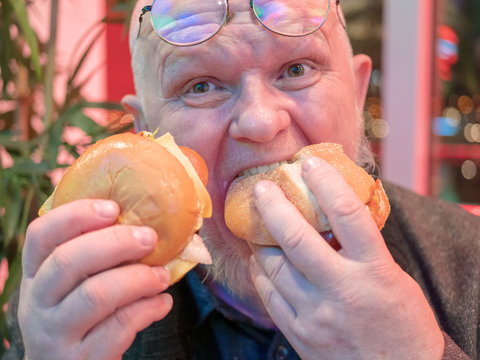 elderly pensioner in fashionable jacket with gray beard in glasses holds hamburgers in two hands. old hungry man, hungry and angry, bites off hamburger alternately from each hand. Fast food cafe