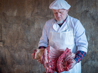 elderly man with a gray beard in cook suit and white apron cuts into pieces large piece of pork with knife and ax
