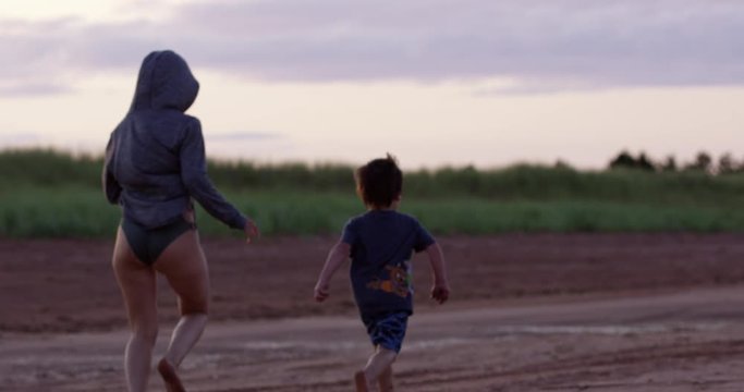 Young mother and toddler boy running on beach during sunset as it begins getting darker and cold