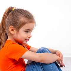 Portrait of a  little smiling girl wearing orange shirt and jeans sitting on the floor clasping her...