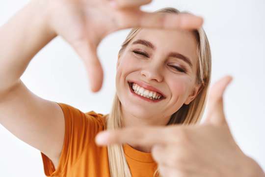 Happy, carefree lovely young blond woman feeling delighted and joyful, close eyes and smiling cheerful, make finger frames as searching for perfect shot, inspirational photo, white background