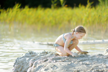 Fototapeta premium Young girl taking healing mud baths on lake Gela near Vilnius, Lithuania. Child having fun with mud. Kid playing with clay.