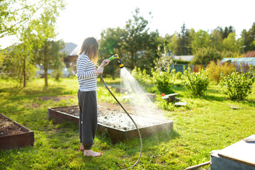 Cute young girl watering flower beds in the garden at summer day. Child using garden hose on sunny...
