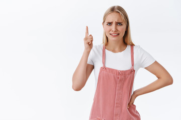 Confused and skeptical young blond girl, cringe face and smirk doubtful, frowning from dislike, pointing up and look camera, showing something strange and disgusting, white background