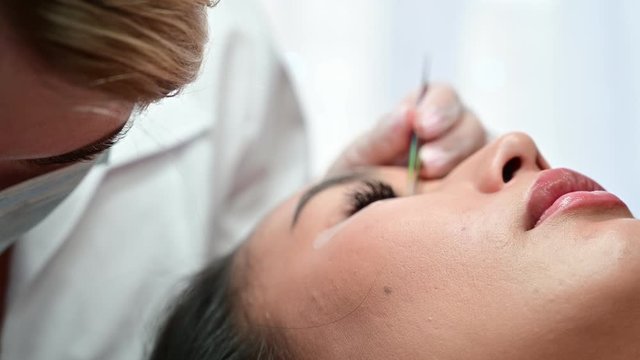 Close-up of a beautiful asian woman on eyelash extension procedure. Double volume false eyelashes. Lashmaker at work.