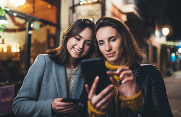 Bloggers together point finger on screen smartphone on background bokeh light in night city, smile girls friends using in hands mobile phone closeup, online wi-fi internet