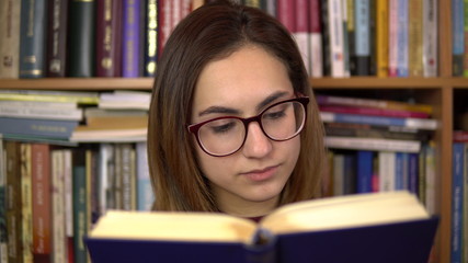 A young woman is reading a book in a library. A woman with glasses carefully looks at the book closeup. In the background are books on bookshelves. Book library.