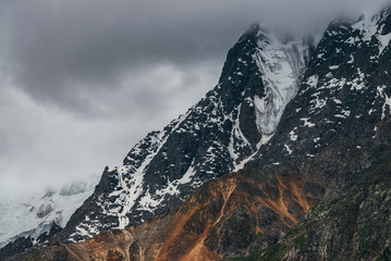Low stormy clouds touch top of snowy mountain. Dismal overcast awesome landscape with big rocks and glacier. Gloomy atmospheric highland scenery in bad cloudy weather. Storm is coming due to mountains