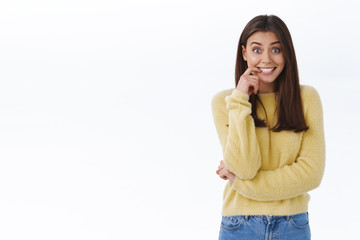 Fototapeta premium Excited and upbeat young feminine brunette woman in yellow sweater smiling thrilled and overwhelmed, biting finger as looking hopeful camera, anticiapting good news, stand white background curious