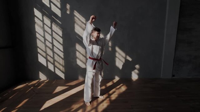A young athlete is preparing for a taekwondo fight. White kimono with a red belt, barefoot, sunlight from the window, training room. camera approaching and zoom in to boy.