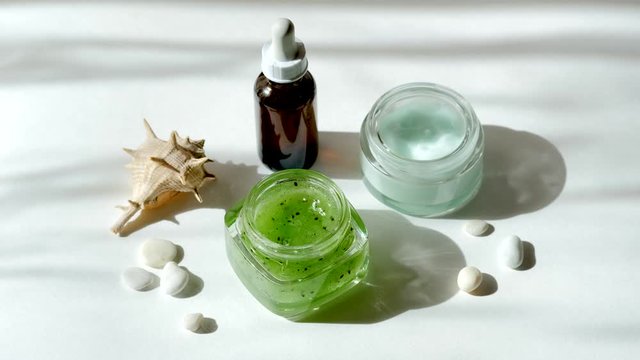 Modern skincare concept. Glass jars with natural green kiwi fruits scrub, moisturizing cream, bottle of hyaluronic acid or cosmetic serum and sea shell in morning sun rays. Natural SPA cosmetic set. 