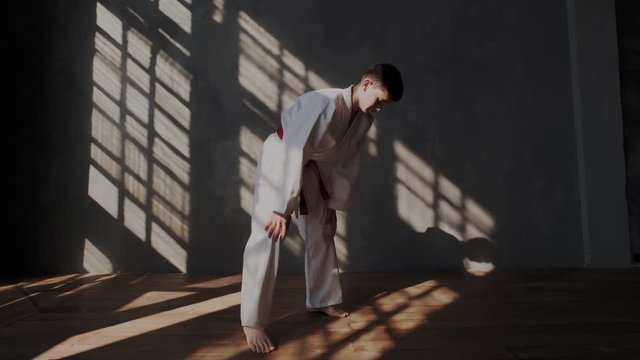 Male brave athlete is preparing for a taekwondo fight. White kimono, barefoot, sunlight from the window, solar grid on the wall and floor. Training room. Camera approaching to boy.