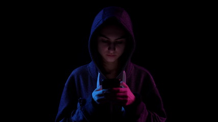 A young woman in a hood with a phone in her hands. Hacker makes a hack through the phone. Blue and red light falls on a person on a black background. © Vital9c