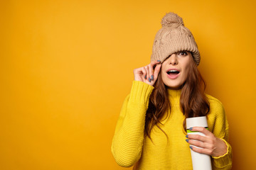A brunette in a yellow sweater with a thermos is standing on a yellow background, pulling a hat over face, mouth open in surprise