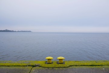 Obraz premium Two yellow mooring bollards on empty dock by the lake off season in misty day. Kamien Pomorski, Poland