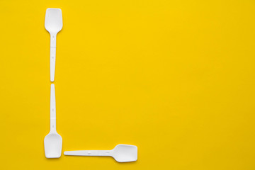 White plastic spoons in the form of letter L on yellow background. Top view. Copy, empty space for text
