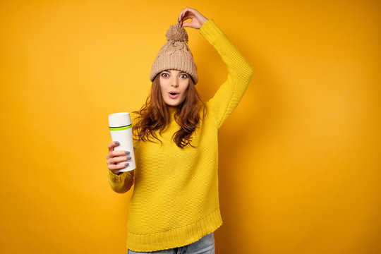 A brunette in a yellow sweater with a white thermos stands on a yellow background and pulls her cap up by the ponpon