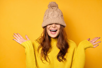 A girl in a yellow sweater stands on a yellow background with a cap pulled over face and laughs, spreading arms out to the sides