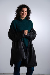 Portrait of happy brunette woman in sweater and leather jacket. Smiling female curly haired model looking at camera. Gray background.