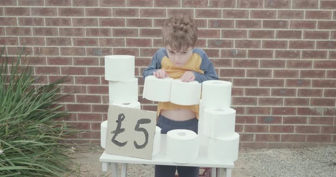 Little boy has a toilet paper stand outside of his house selling rolls of toilet paper taking advantage of a toilet paper shortage. Hand painted sign with five pounds written on it