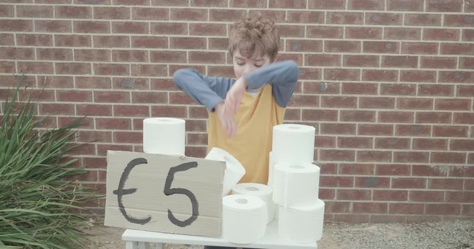 Little boy has a toilet paper stand outside of his house selling rolls of toilet paper taking advantage of a toilet paper shortage. There is a hand painted sign with five euros written on it