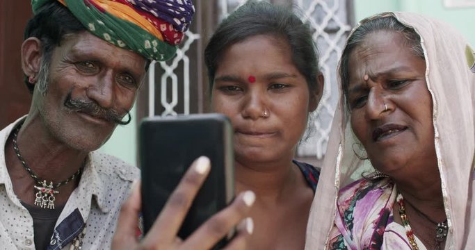 Confident young teenage woman with her traditional grandparents in Rajasthan, India as she takes selfie photo video on a smart phone mobile touch screen technology device and shares and shows them
