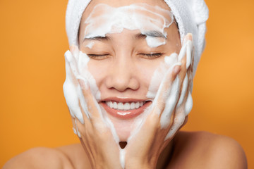 Young Asian woman applying cream on her face and touching face while wearing a bath towel. Isolated...